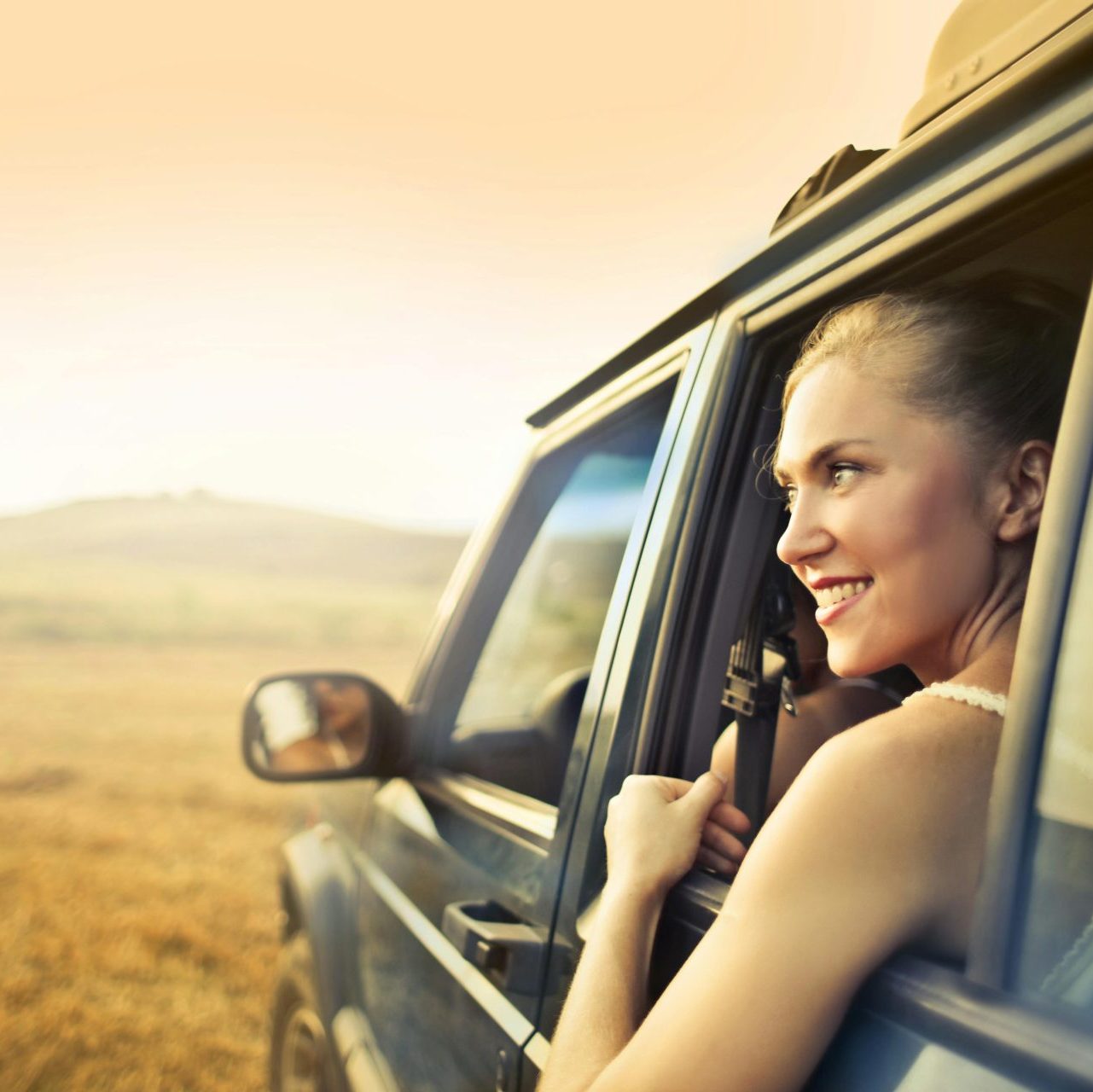 Young happy female smiling and enjoying sunset through open window while traveling by car